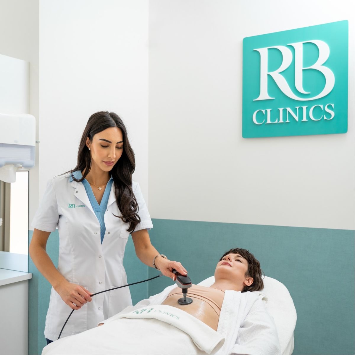 Specialist van RB Clinics behandelt de buik van een cliënte met de Indiba applicator, met het RB Clinics logo aan de wand